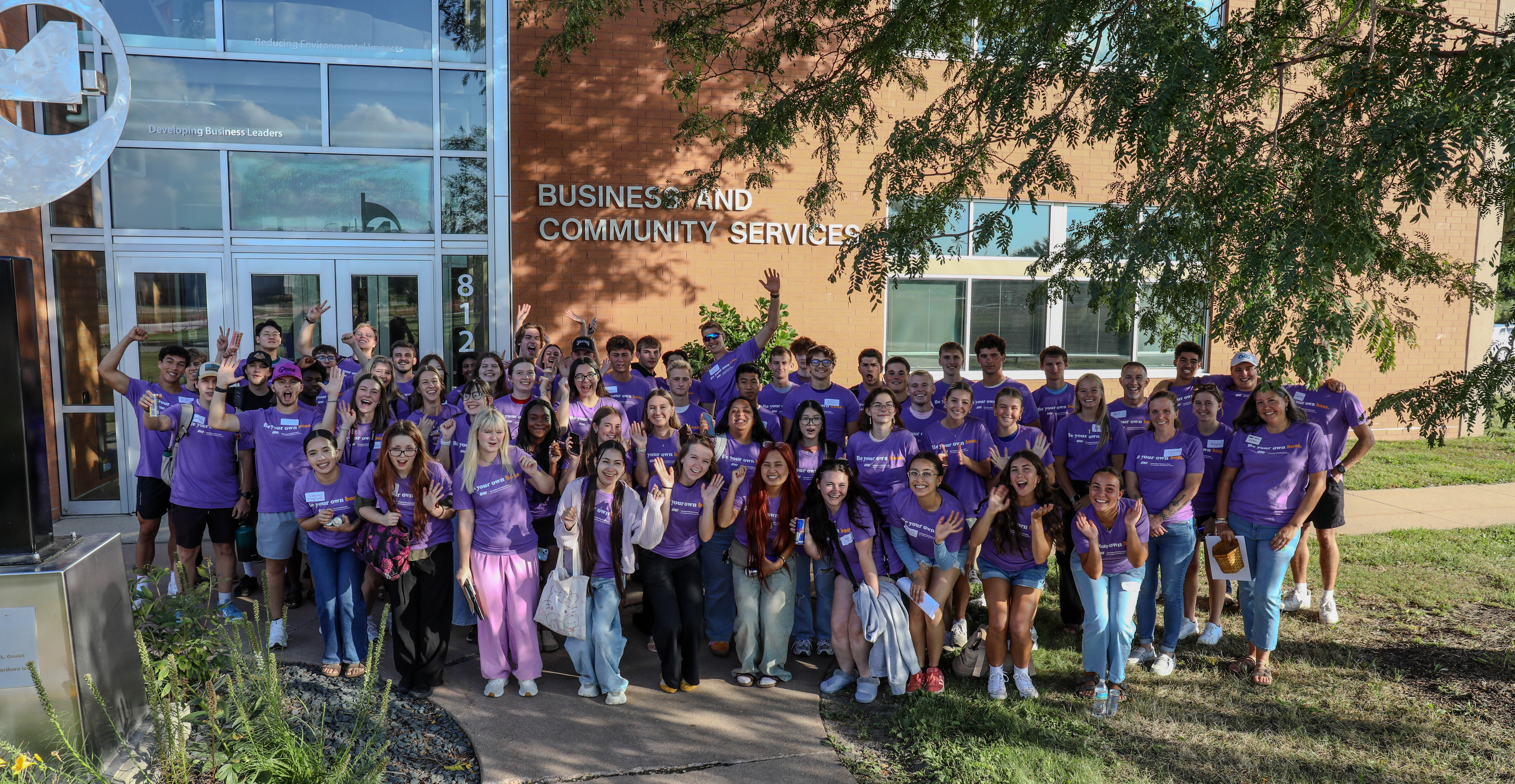 Large group of students in purple 'Be Your Own Boss' T-shirts posing and cheering outside UNI's Business and Community Services building.