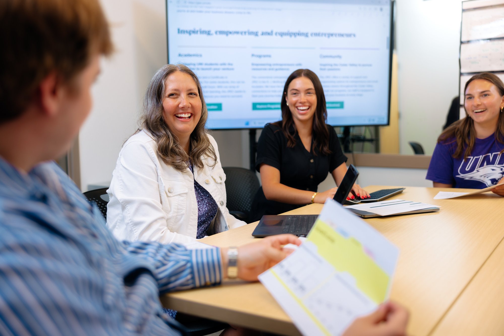 Group of people smiling and collaborating around a table with laptops and papers, with the JPEC website displayed on a screen behind them, one person wearing a UNI shirt