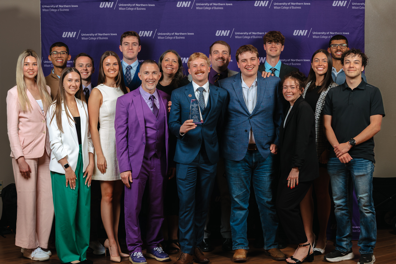 A large group poses together in front of a purple UNI Wilson College of Business backdrop, with one man in a purple suit and another holding a trophy at the center of the group. Everyone is dressed in business or business-casual attire, smiling for the photo.