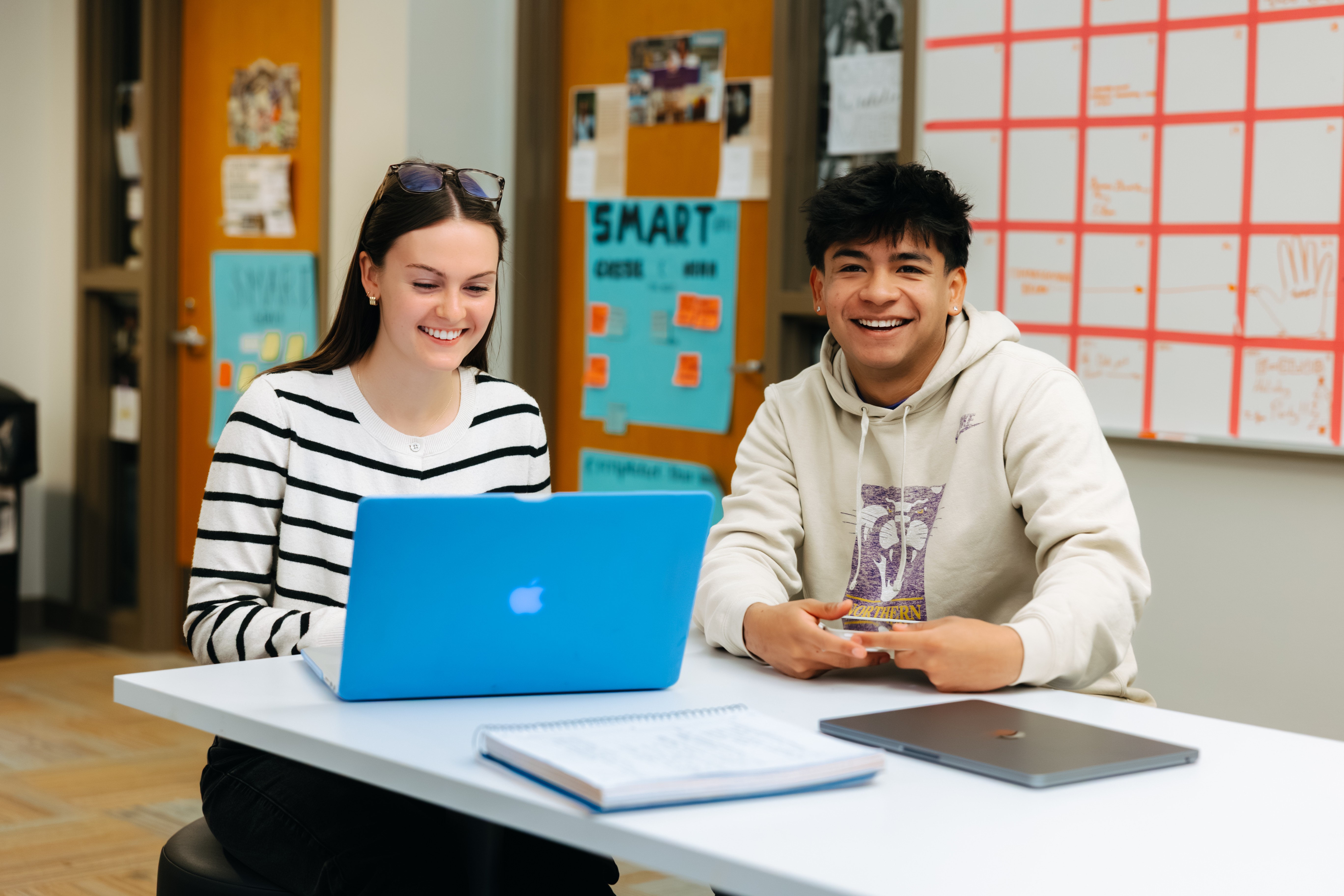 Two students smile while sitting at a table with a blue MacBook, notebook, and laptop.