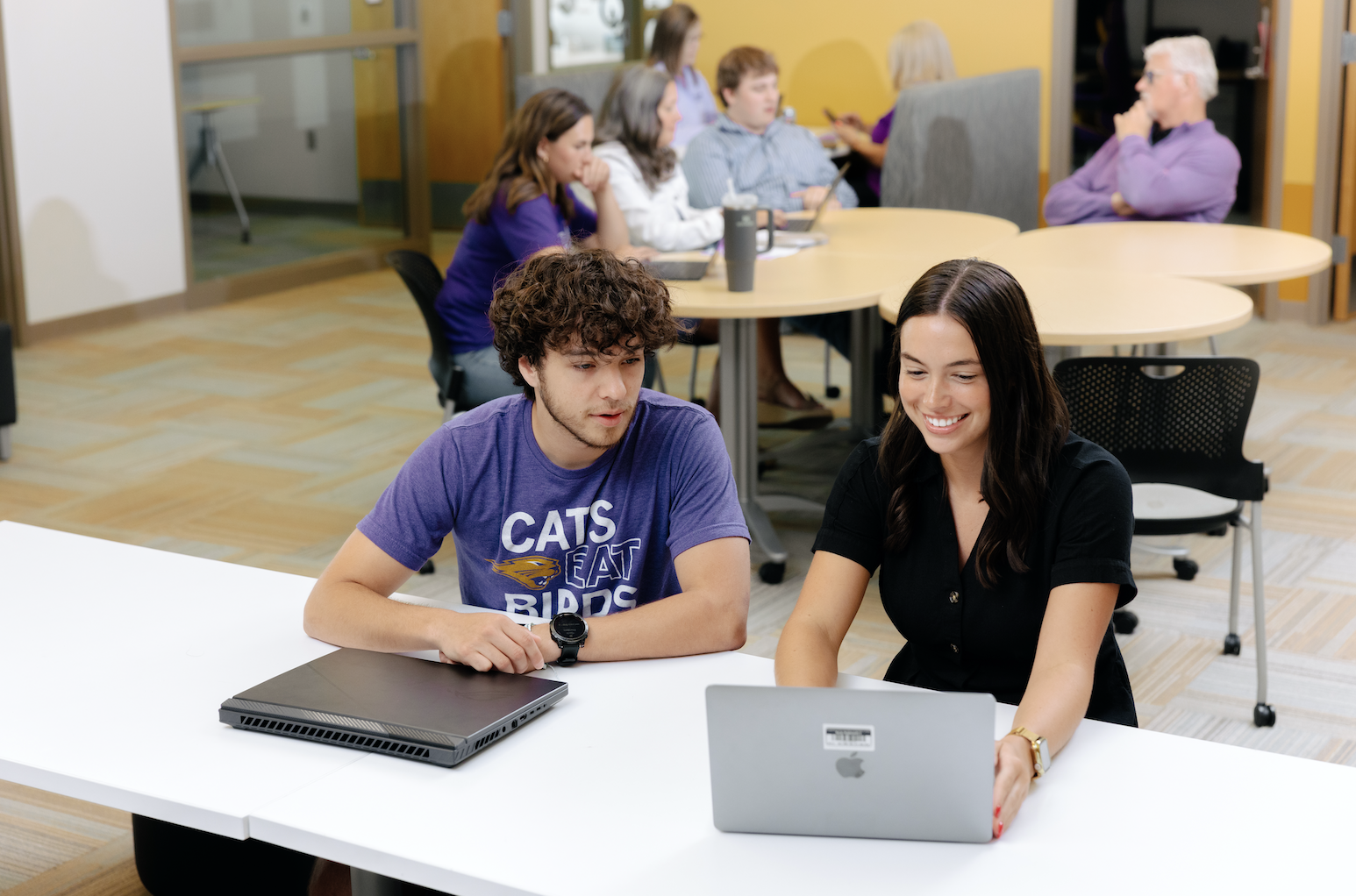 A student in a purple "Cats Eat Birds" t-shirt sits at a table with a laptop, talking with a staff member in black who is smiling and gesturing toward her own laptop. In the background, other groups work together at round tables in an open, sunlit space.